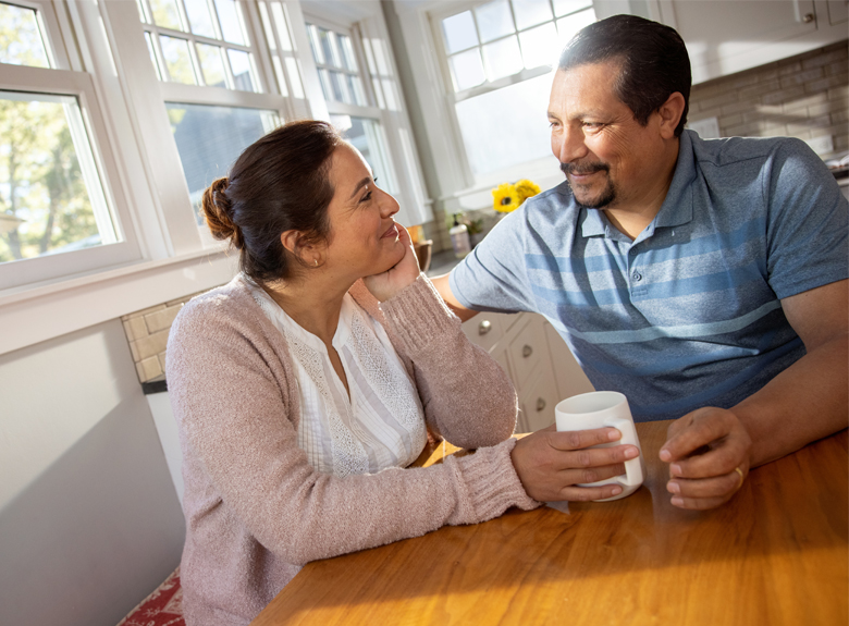 A man and woman sit at a table looking at each other and smiling. The woman is holding a cup.