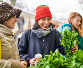 Three women dressed for winter in jackets, scarves, and hats are smiling while outside.