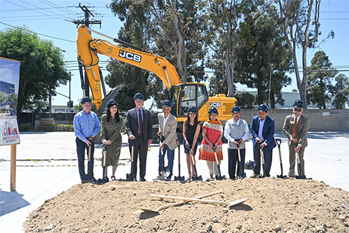 The Shah family stands outside in front of a large piece of construction equipment to break ground together.