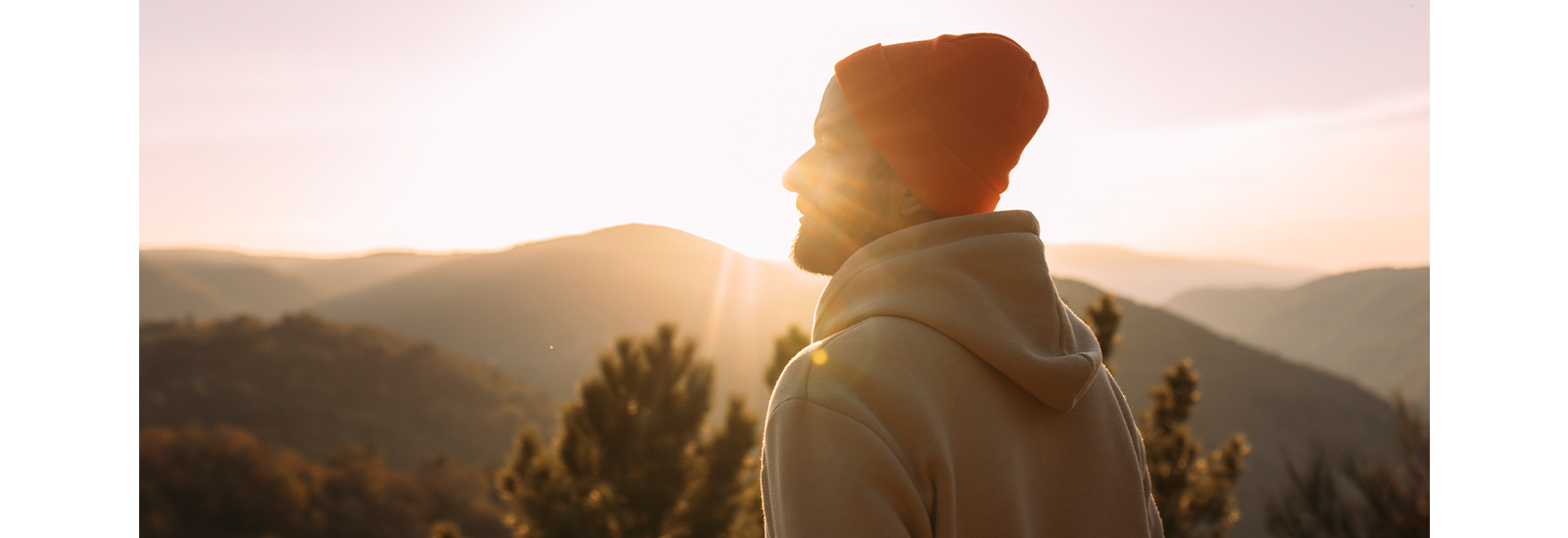 A man in a hoodie and ski cap looks toward the horizon as the sun rises