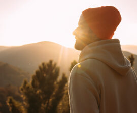 A man in a hoodie and ski cap looks toward the horizon as the sun rises