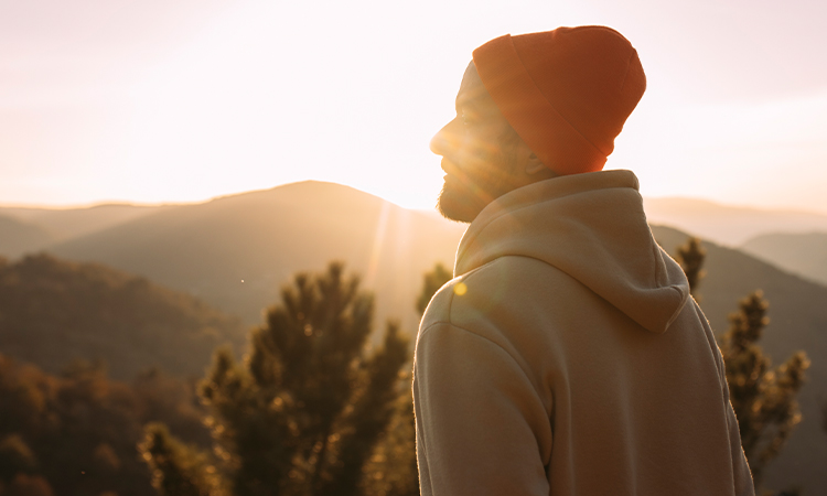A man in a hoodie and ski cap looks toward the horizon as the sun rises