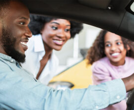 A man sits in a car as his wife and daughter look through the driver’s side window