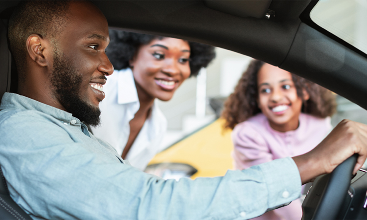A man sits in a car as his wife and daughter look through the driver’s side window