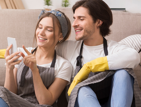 A man and woman wearing aprons and gloves sit in front of a couch looking at a phone.