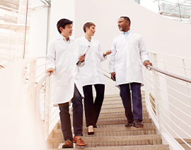 Three people in white coats stand on a stairway talking.