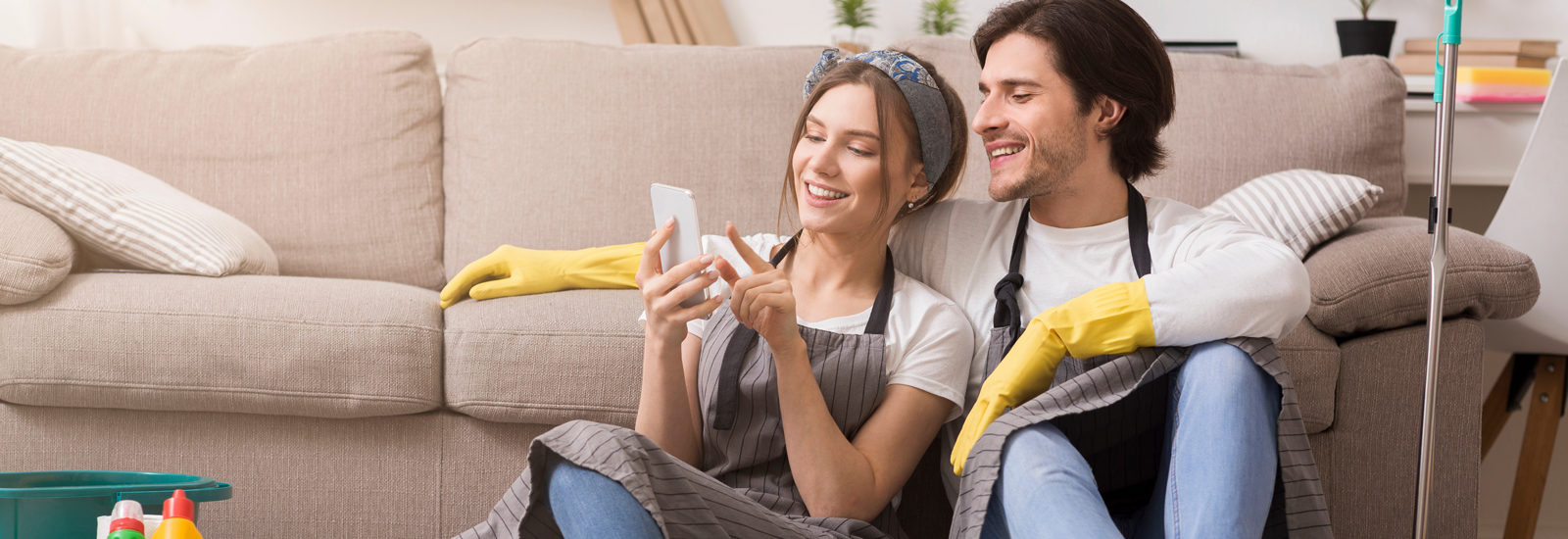 A man and woman wearing aprons and gloves sit in front of a couch looking at a phone.