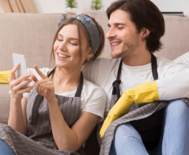 A man and woman wearing aprons and gloves sit in front of a couch looking at a phone.