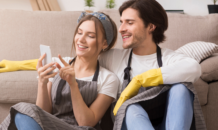 A man and woman wearing aprons and gloves sit in front of a couch looking at a phone.