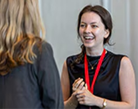 Two individuals are talking - one has her back to the camera. The one speaking wears a red lanyard and has her hands clasped.