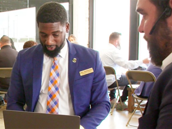 A man in a suit sits at a table looking at his laptop