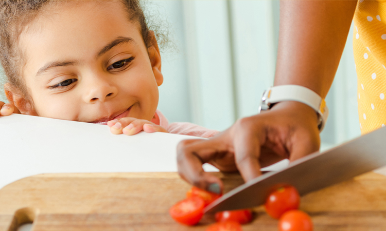 A young child watches as her parent cuts tomatoes.