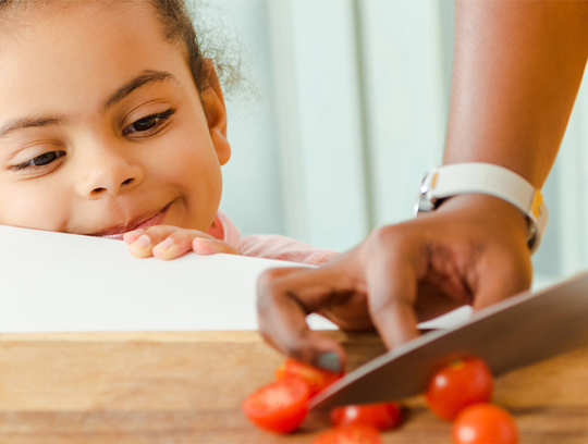 A child peers over the counter as an adult chops a pepper.
