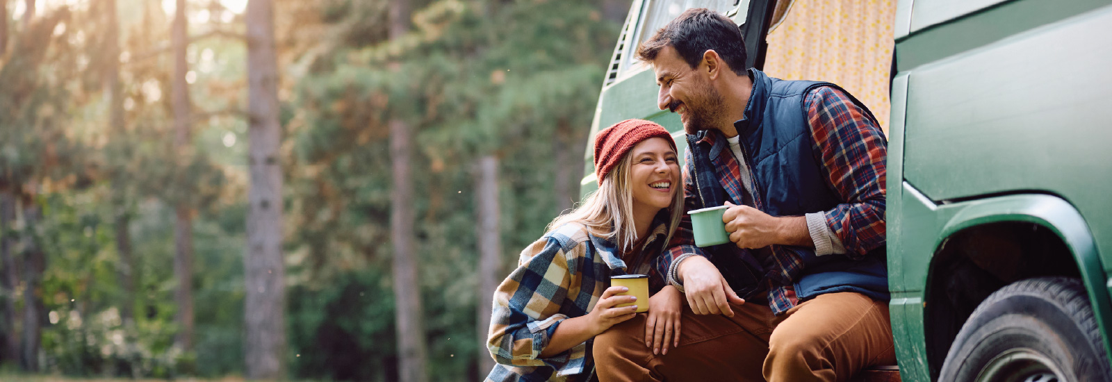 Two adults sit beside a recreational vehicle in a wooded area.