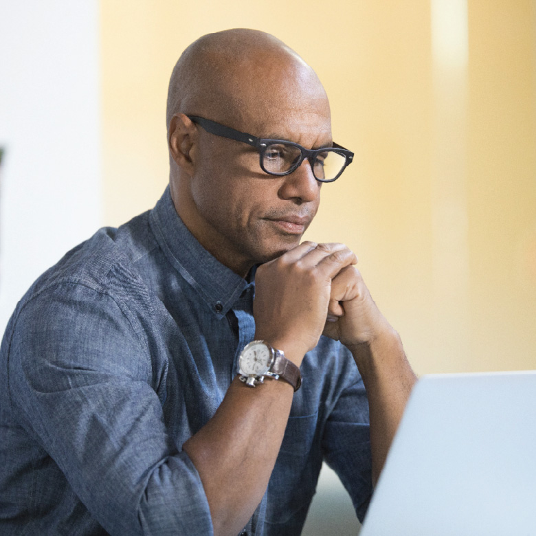A man wearing glasses and a blue shirt is seated in front of an open laptop, focused in thought.