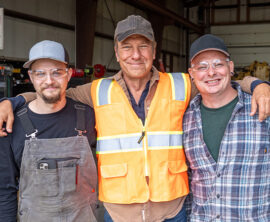 Three smiling men stand together in a workshop