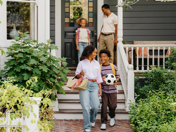 A family is walking down their front steps. The children are in backpacks and the son holds a soccer ball.