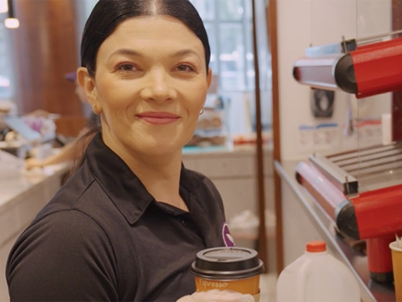 A woman holding a coffee cup, working in a cafe, smiles at the camera.