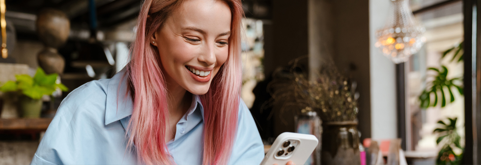 Person with pink hair holding a smartphone while seated in a café-like setting with plants and decorative lighting in the background.