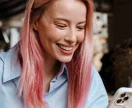 Person with pink hair holding a smartphone while seated in a café-like setting with plants and decorative lighting in the background.