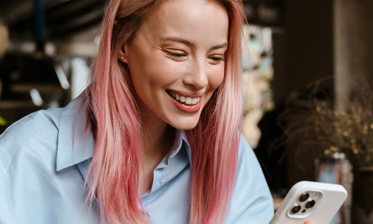 Person with pink hair holding a smartphone while seated in a café-like setting with plants and decorative lighting in the background.