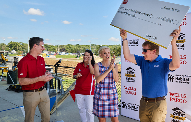 Four people stand outdoors near a construction site while one person raises an oversized grant check.