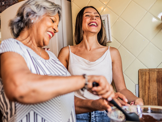 Two women are laughing in a kitchen as they stir ingredients in a bowl.