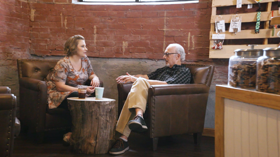 Lindsey Braciale sits on a leather couch with a man. They look at each other while talking.