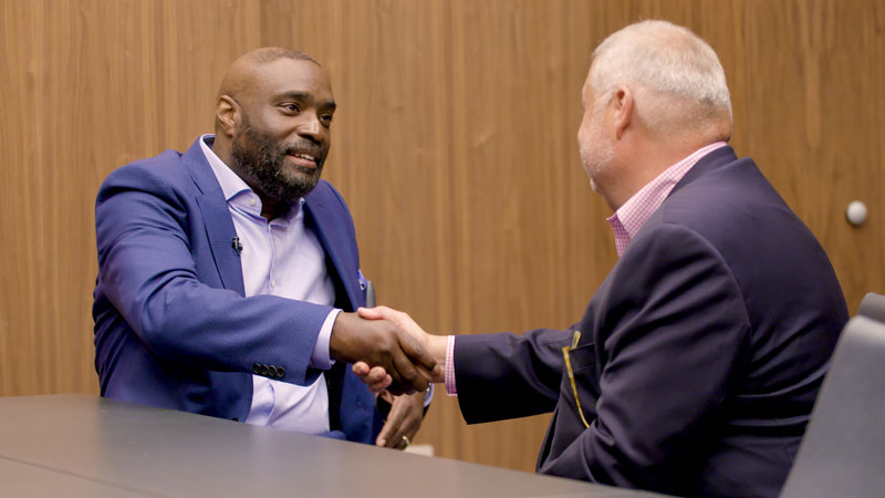 Seated at a conference table, Antwone Fisher and Jon Campbell shake hands at a panel on housing affordability.