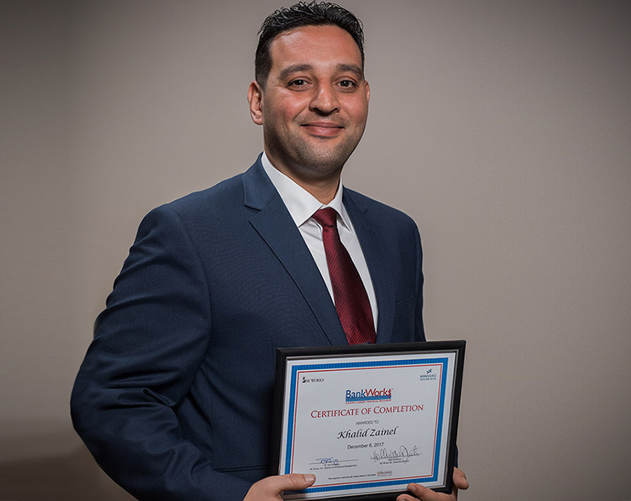 Khalid Zainel wears a suit and smiles as he holds a framed certificate of completion with BankWork$ at the top.