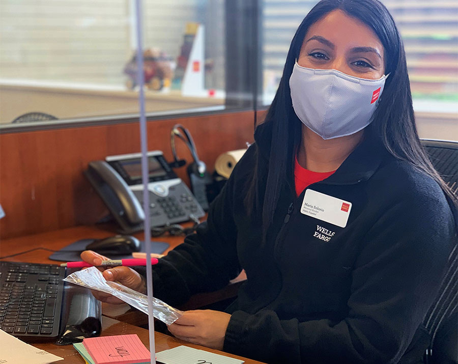 Maria Solorio sits at a desk inside a bank branch while wearing a mask and looking ahead. Her mask, black coat, and nametag all have Wells Fargo’s logo on them.