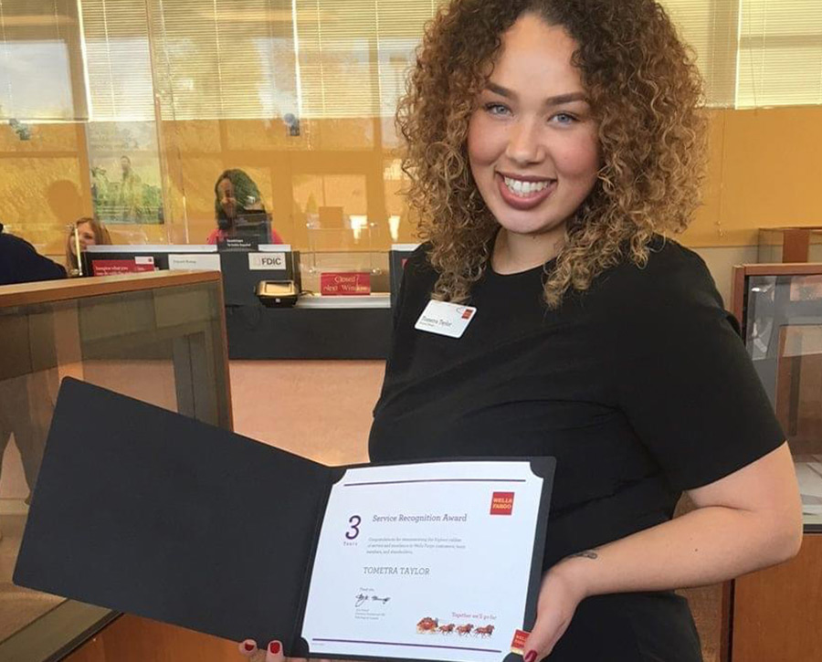 Tometra Taylor stands inside a bank branch and smiles while holding an open folder. Inside is a paper with a 3, indicating years of service, and Wells Fargo’s logo.
