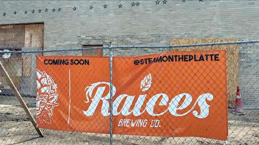 An orange sign for Raices Brewing Co. hangs on a chain link fence outside a concrete warehouse.