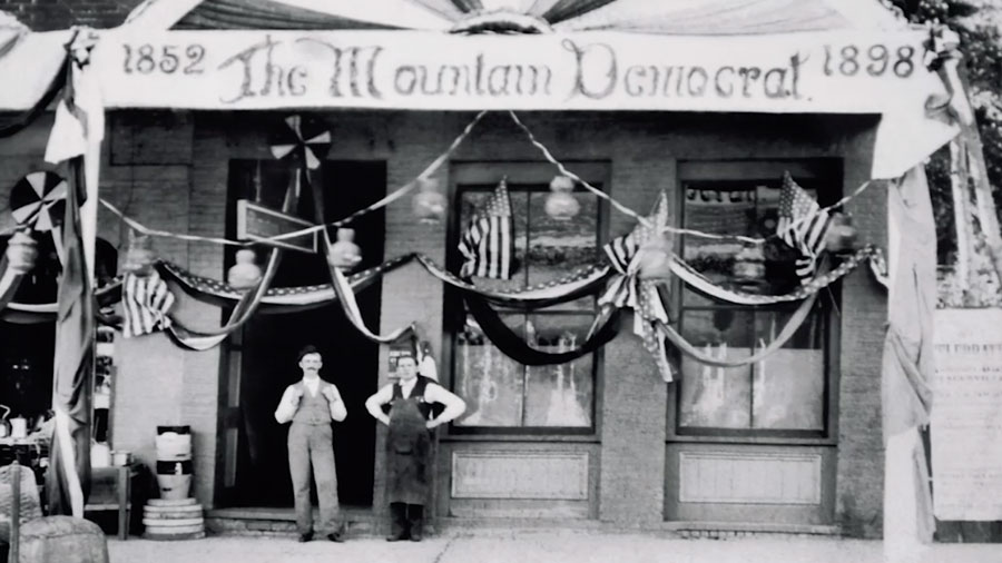 In a very old photo, two men stand outside of a building decorated with American flags and streamers. A banner above the men reads, “The Mountain Democrat” and the dates “1852” and “1898.”