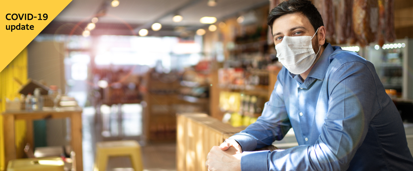 A business owner, wearing a blues dress shirt and a face mask, stands in his restaurant and food market. A graphic on the image says 'COVID-19 update.'
