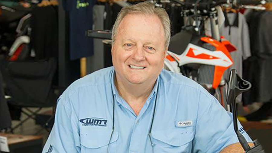 In a light blue shirt bearing his company’s logo, Bob Brewster, owner of WMR Competition Performance, smiles in his dealership showroom with dirt bikes in the background.