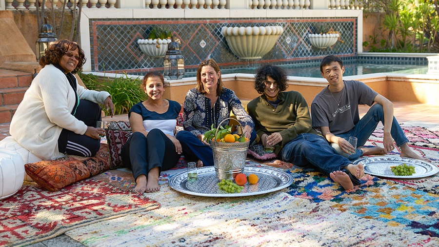 A group of five people sit on carpets outside and smile ahead. There are several silver platters and a silver container with food in front of them.