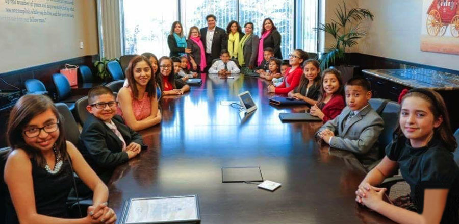 Los niños están sentados alrededor de una mesa en una sala de juntas en la sucursal de Wells Fargo en Fresno, California. Los adultos están de pie al fondo de la sala.