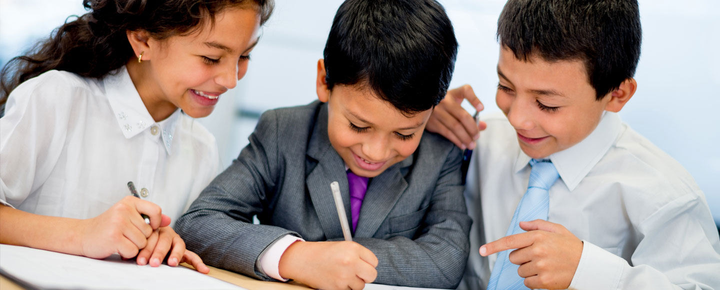 Una niña y un niño sonríen al niño sentado entre ellos mientras escribe números en una hoja de papel durante una lección sobre finanzas personales.