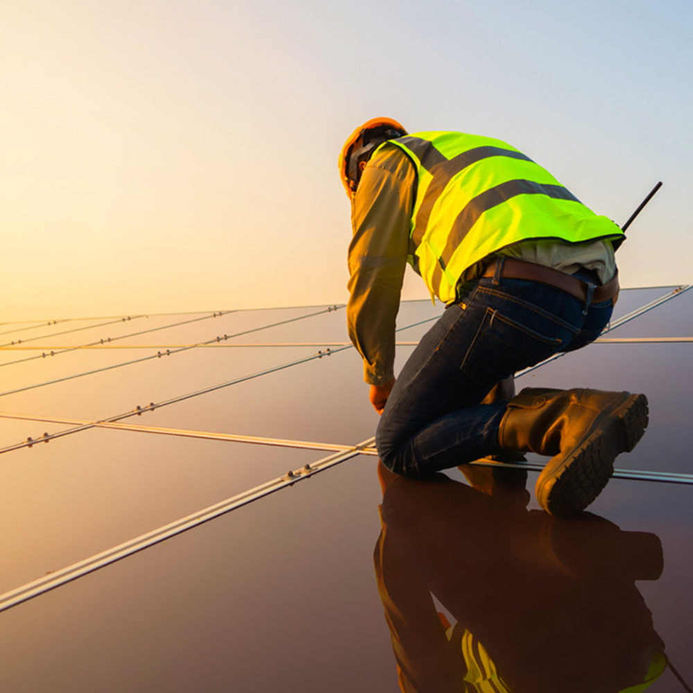 A man wearing a hard hat and safety vest kneels down to work on a rooftop solar panel.
