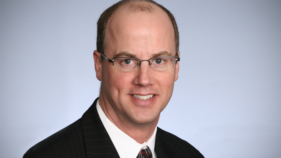 The headshot of John Rasmussen shows him smiling at the camera while wearing glasses, a dark blazer and tie, and a white, collared shirt.