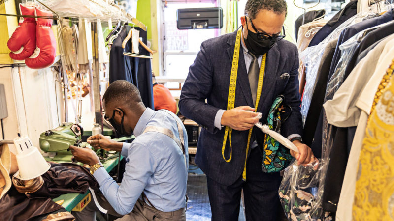 Eddie Lofton, wearing a suit with a face mask and a measuring tape around his neck, cinches a garment bag while one of his employees sits at a sewing machine on his right.