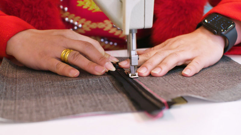 A closeup of a woman’s hands sewing a zipper to fabric with a sewing machine.