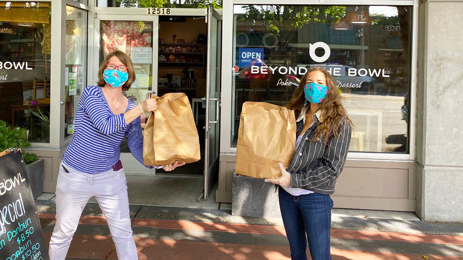 Two Build Lake City Together volunteers hold grocery bags of meals bought from the Beyond the Bowl restaurant behind them for the 