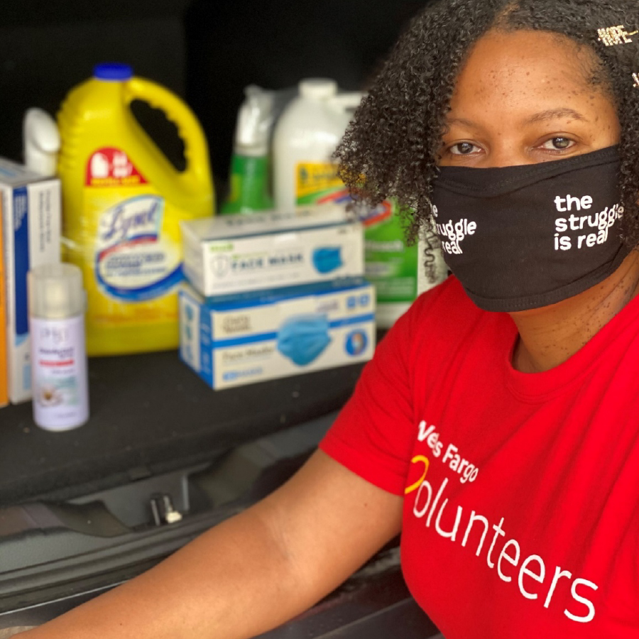 A woman looks at the camera while she wears a red shirt with “Wells Fargo volunteers” on it and a black mask saying, “The struggle is real.” Behind her are various bottles and boxes that are out of focus.