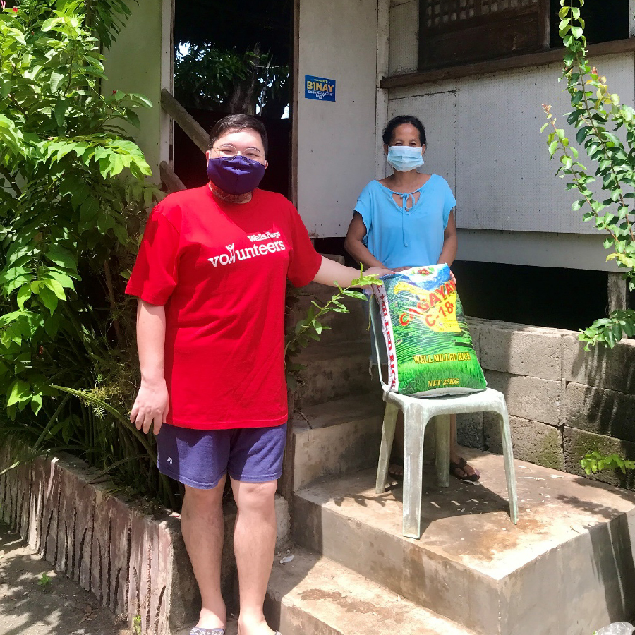 A man wearing a red shirt and mask looks at the camera while holding a bag of rice. The bag is on a chair in front of a woman who also wears a mask and stands nearby on some steps.