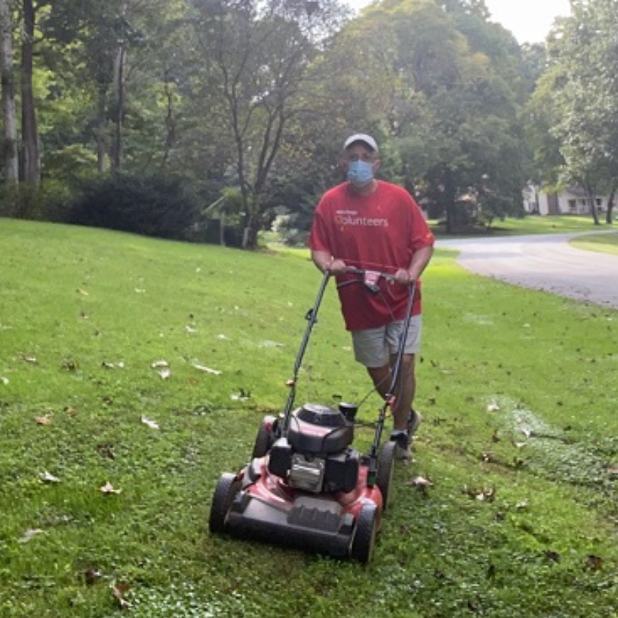 A man wearing a mask and red shirt uses a walking lawn mower.