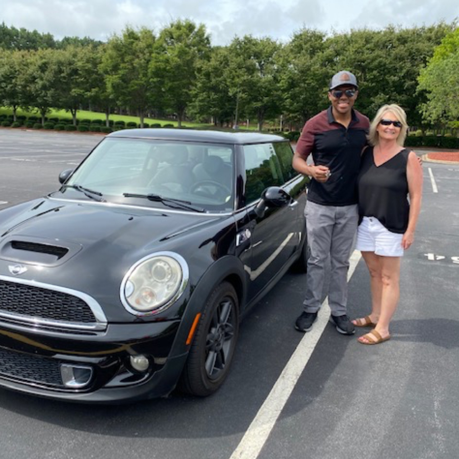 A man and woman stand next to a car parked outside and smile at the camera.