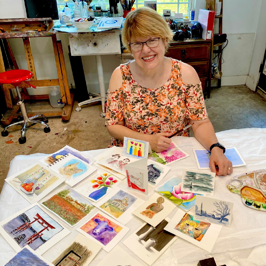 A woman sits at a table with postcards on it and smiles at the camera. The postcards have paintings on them.