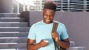 Photo of a young college student, standing on the steps of a library or other campus building, holding his phone and smiling.
